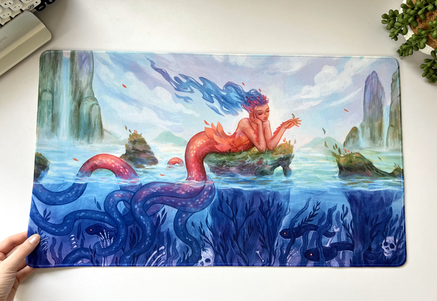 A beautiful mermaid lounges on a mossy rock in the middle of a lagoon. But beneath the water are bones and deep sea creatures.