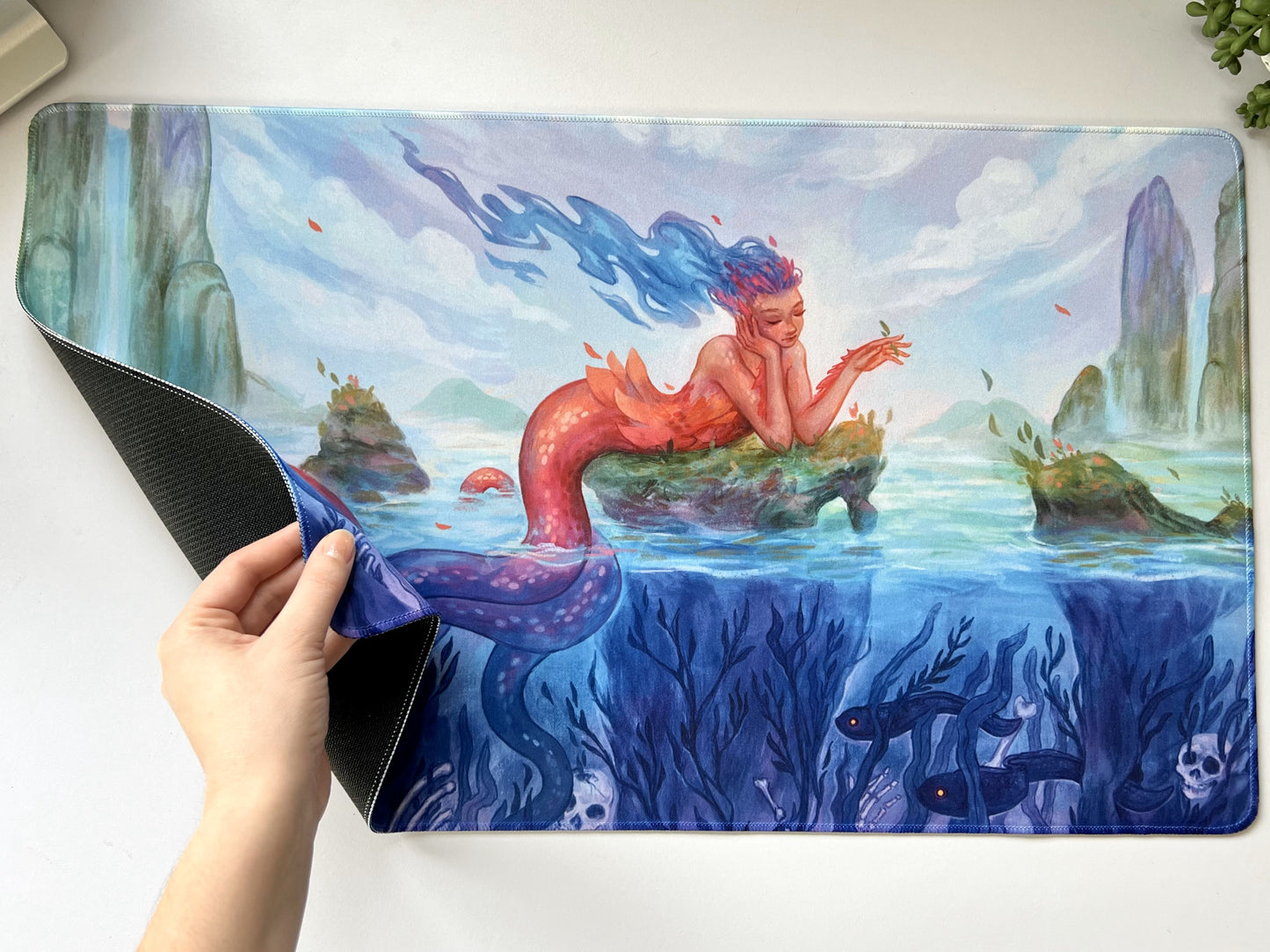 A beautiful mermaid lounges on a mossy rock in the middle of a lagoon. But beneath the water are bones and deep sea creatures.
