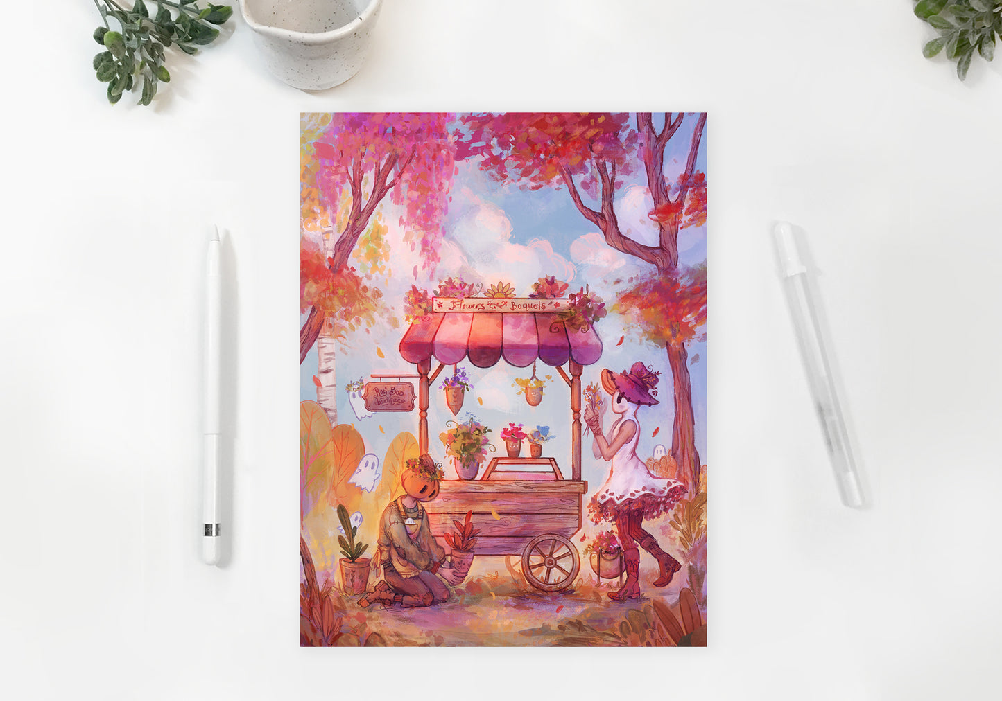 A flatlay of the print on a white table. green leaves and pens are scattered around the print. On the print, Mr. Pumpkin kneels down caring for a plant. His girlfriend Rosi Boo the ghost girl stands beside her flower cart.