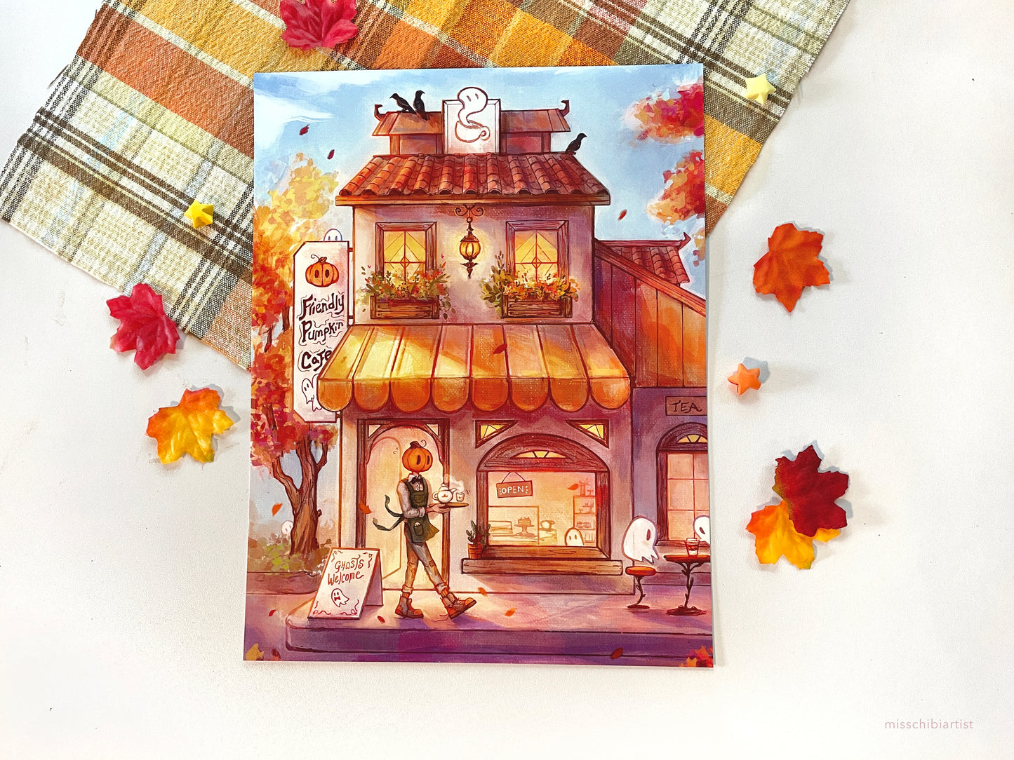A flatlay of the print over a fall plaid cloth. Leaves are scattered around the print. On the print, Mr. Pumpkin carries a tray of tea to some ghosts sitting at an outdoor table. A building in the back says Friendly Pumpkin Cafe.