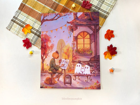 A flatlay of the print over a fall plaid cloth. Leaves are scattered around the print. On the print, Mr. Pumpkin sits outside on the sidewalk painting a ghost family.