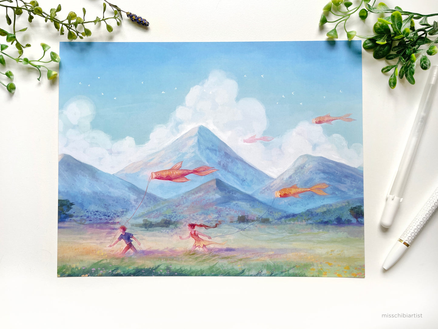 The Carefree illustration shows two children running across a peaceful meadow, holding orange fish kites. The girl has a yellow dress and long brown hair and the boy has short brown hair and a blue shirt. Tall blue mountains rise up in the distance. Available as an art print by MissChibiArist.