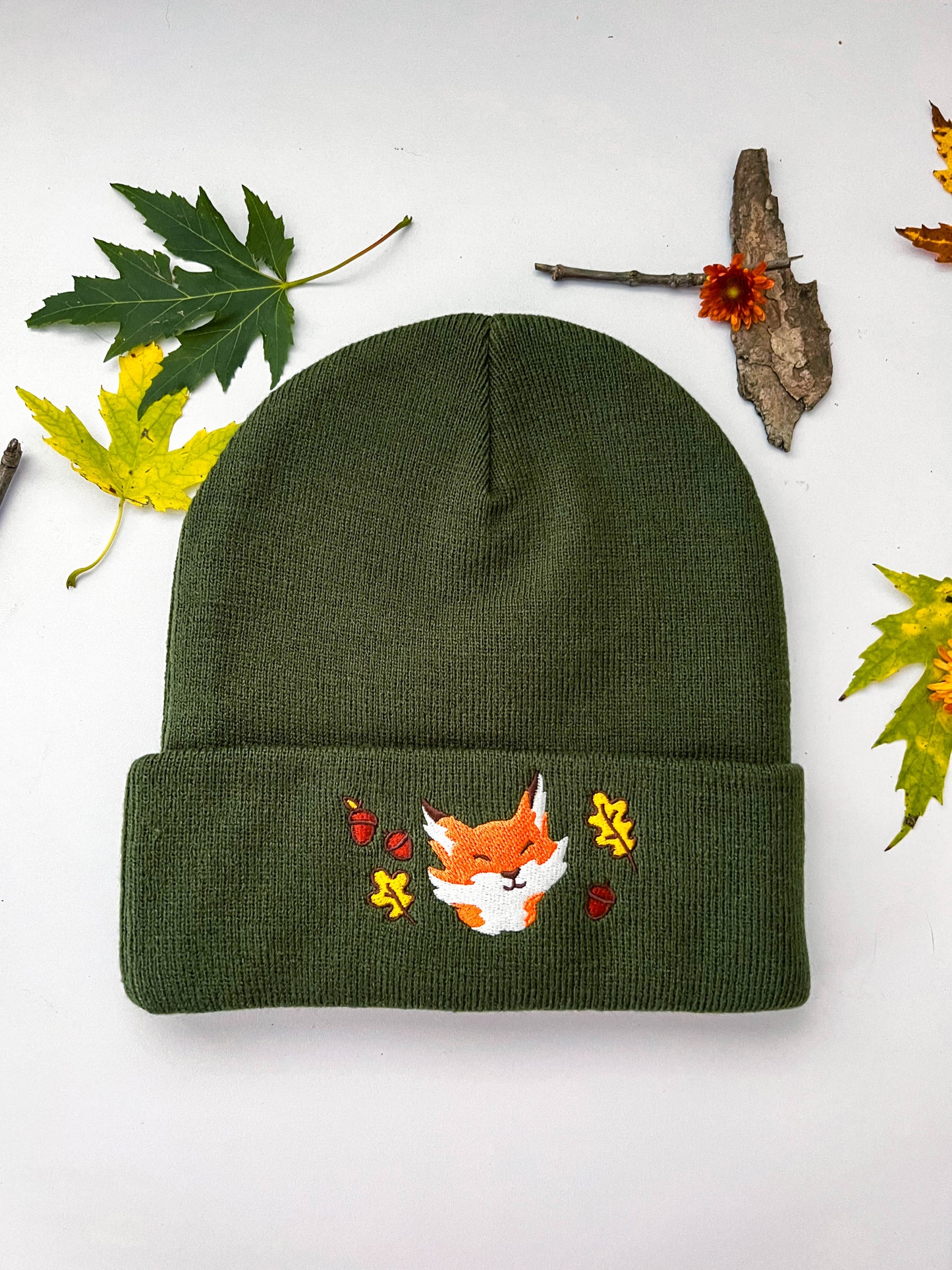 A front view of the forest green fox beanie that shows the embroidery. The portrait of an orange and white fox is smiling with slitted happy eyes. The fox is surrounded by yellow leaves and brown acorns.