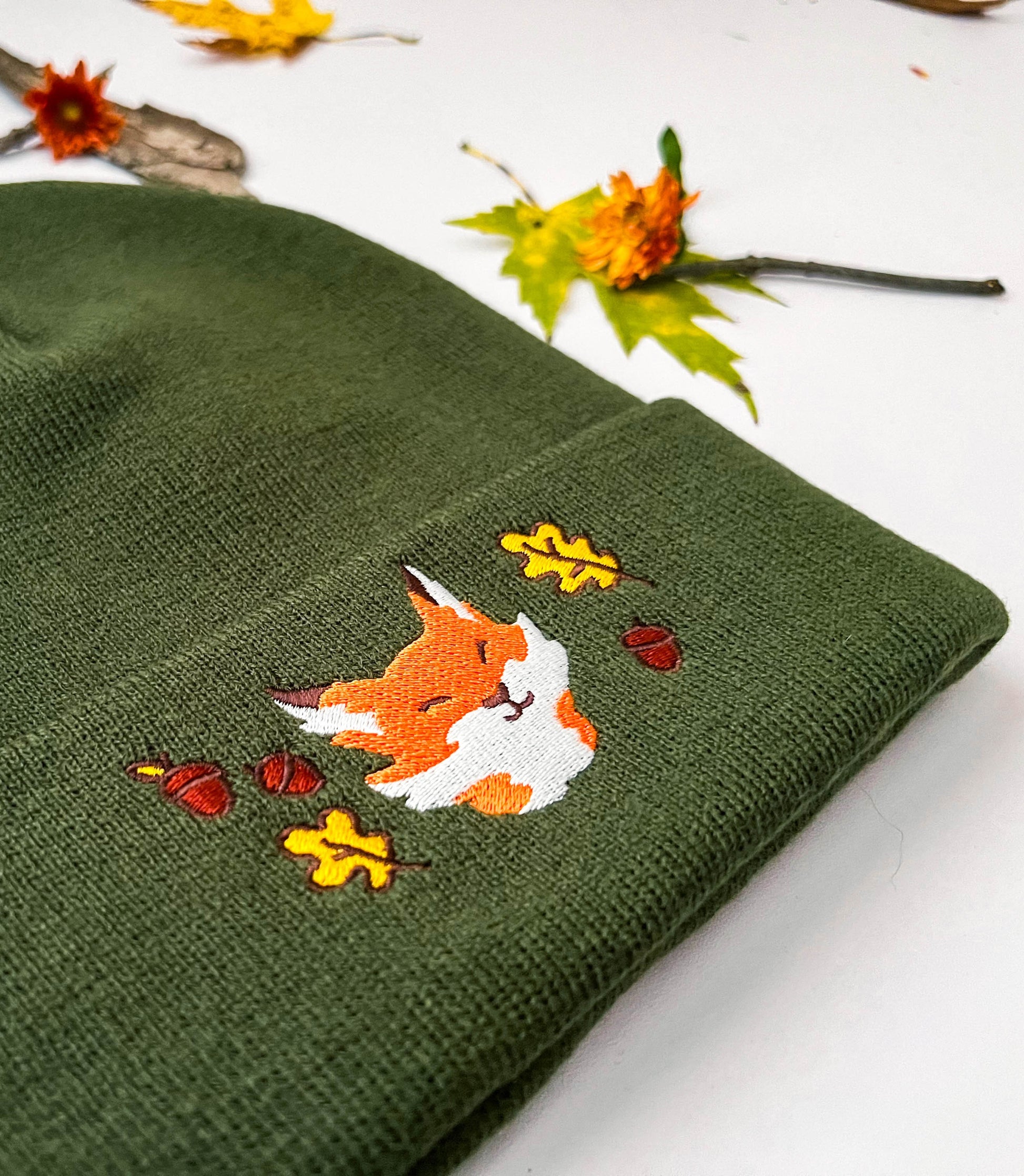 A close image of the forest green fox beanie that shows the embroidery. The portrait of an orange and white fox is smiling with slitted happy eyes. The fox is surrounded by yellow leaves and brown acorns.