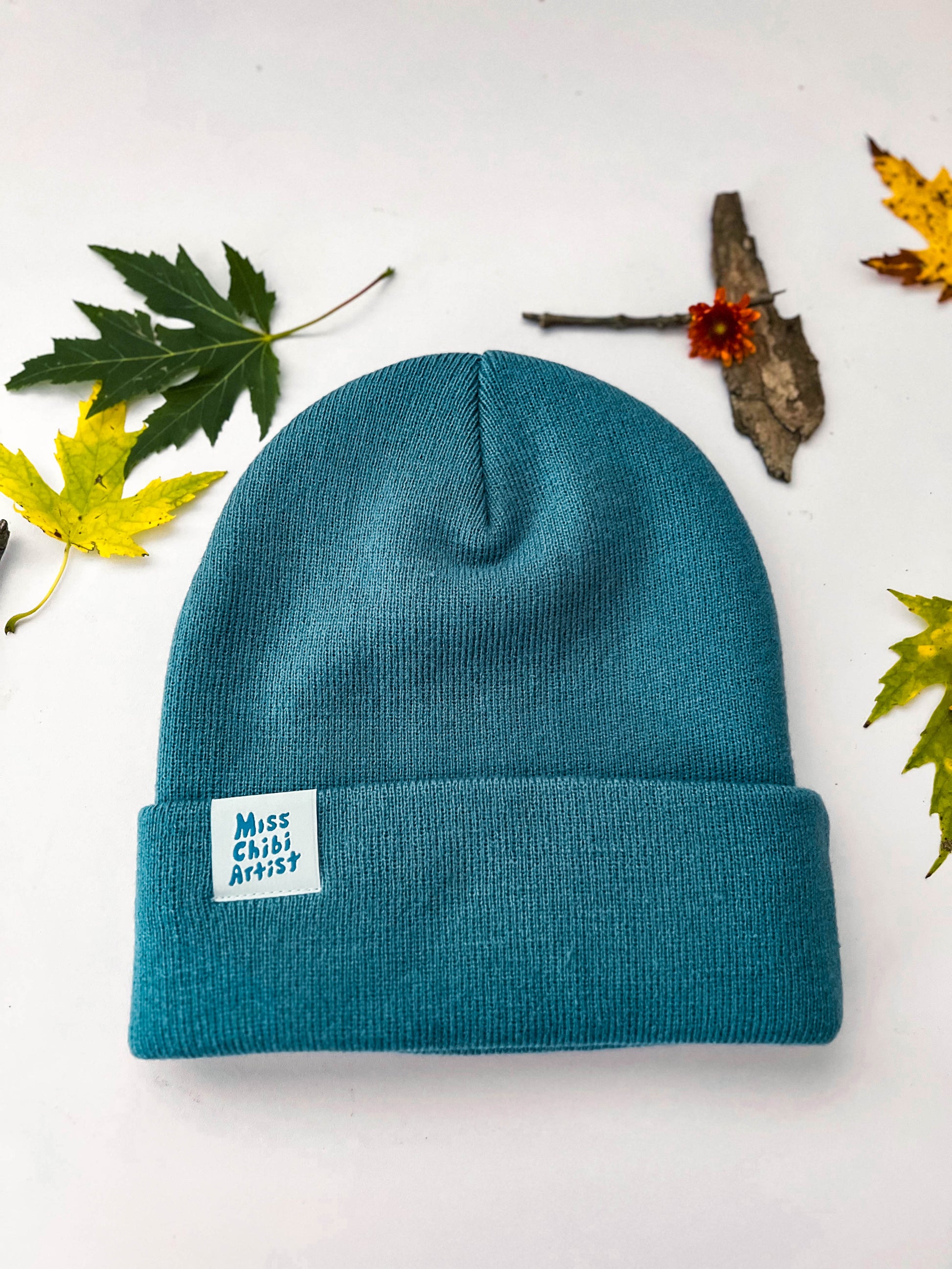 The back of the blue moon beanie has a cream tag that says, "Miss Chibi Artist." Leaves are scattered around the beanie.