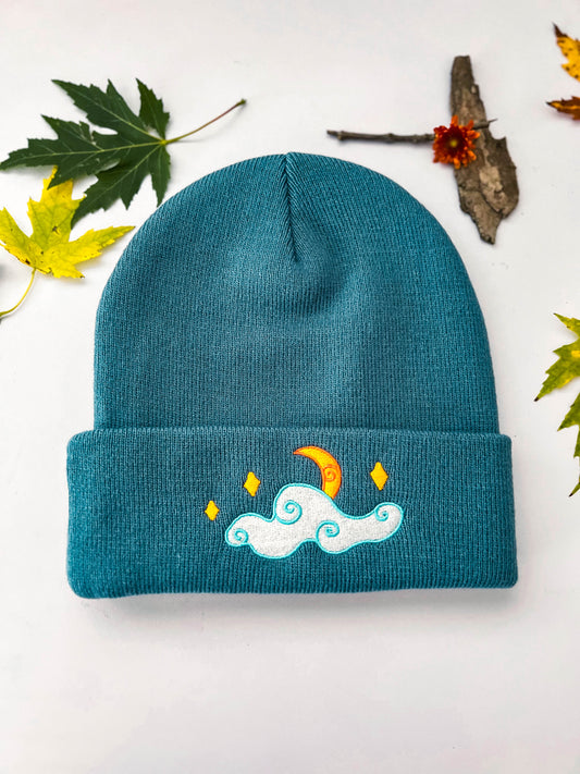 A front view of the blue moon and stars beanie that shows the embroidery. The crescent moon is half-hidden by a cloud with whimsical swirls. Several stars shine above the cloud.