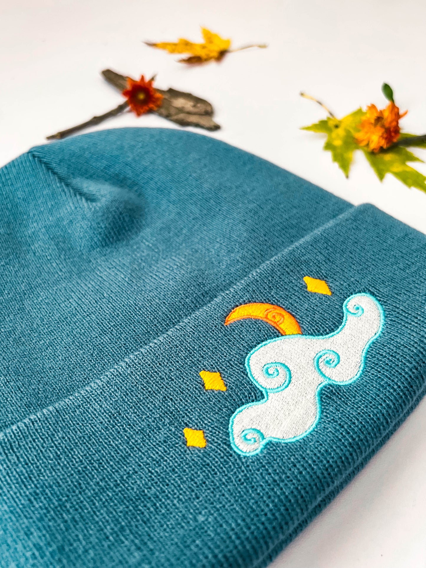 A closer front view of the blue moon and stars beanie that shows the embroidery details. The crescent moon is half-hidden by a cloud with whimsical swirls. Several stars shine above the cloud.