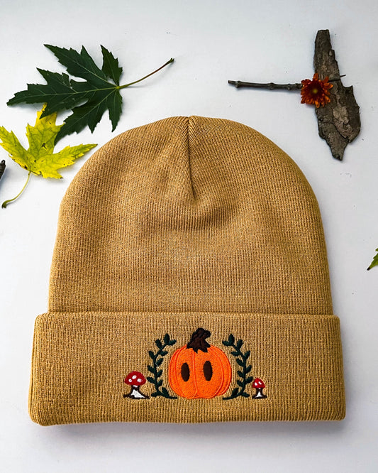 A front view of the light brown Mr. Pumpkin beanie that shows the embroidery. Mr. Pumpkin is an orange and brown pumpkin surrounded by green leaves and red mushrooms.