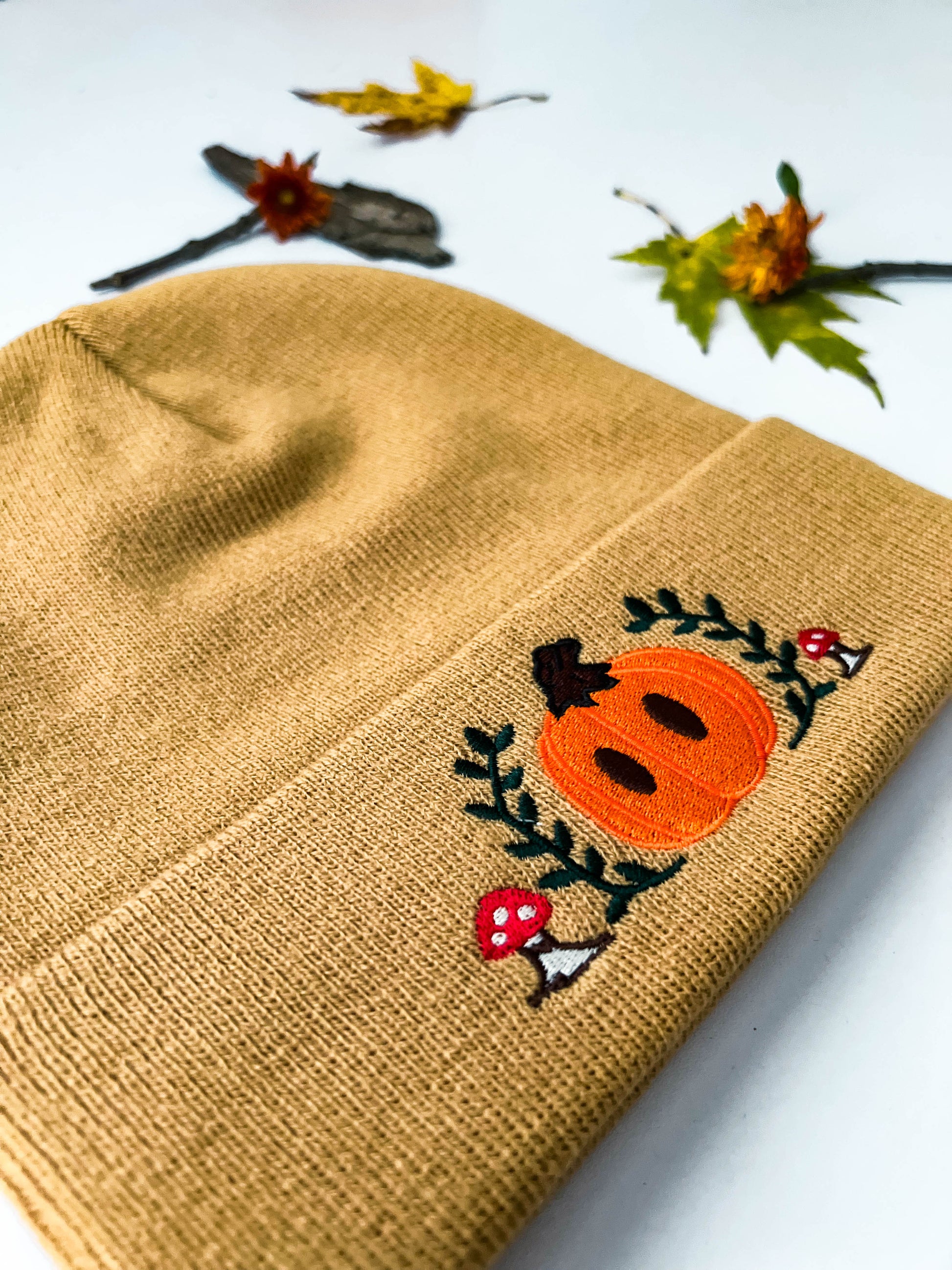 A front view of the light brown Mr. Pumpkin beanie that shows the embroidery. Mr. Pumpkin is an orange and brown pumpkin surrounded by green leaves and red mushrooms.