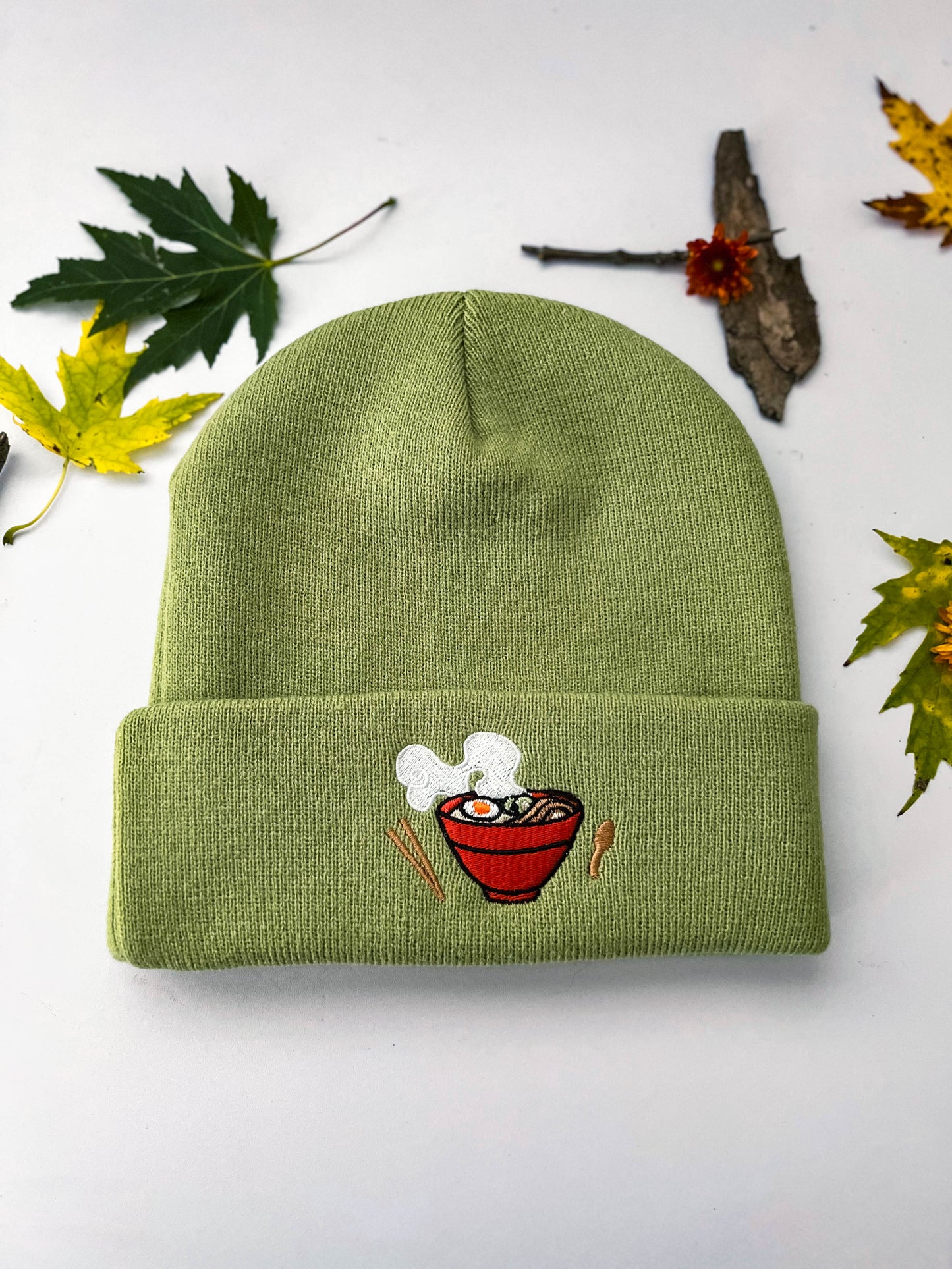 A front view of the light green Ramen beanie that shows the embroidery. There is a red bowl filled with ramen. On each side of the bowl is wooden chopsticks and a spoon.