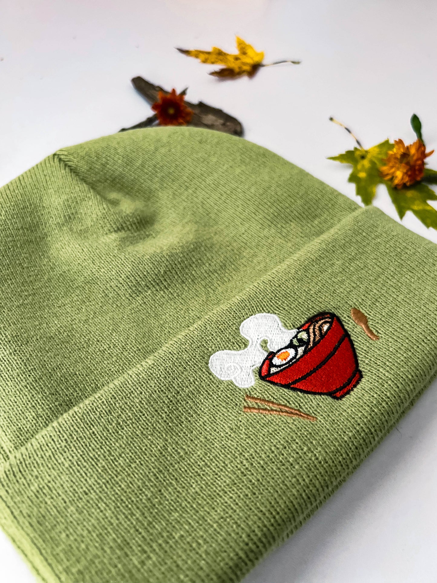 A closer look at the light green ramen beanie that shows the embroidery. There is a red bowl filled with ramen. On each side of the bowl is wooden chopsticks and a spoon.